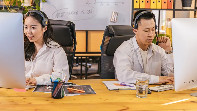 Two office workers with headsets working at computers on a shared wooden desk