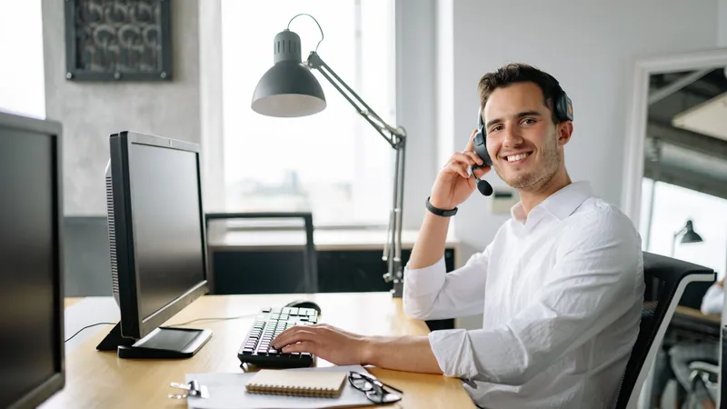 Smiling man wearing a headset making a call at his desk with a computer and notebook