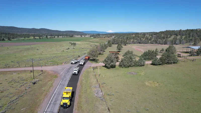 Aerial view of trucks on a two-lane rural road through green fields and pine trees