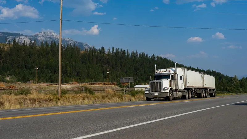 White semi truck pulling two trailers along a highway past a pine forest
